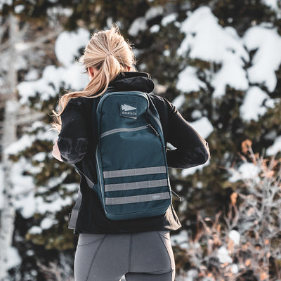 A person wearing a black 15L Bullet Ruck Classic - Cordura and outdoor clothing stands in a snowy forest, their streamlined gear blending seamlessly with the tranquil winter landscape.