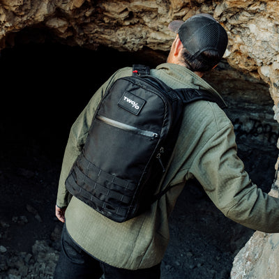 A person in a green jacket and cap, equipped with a Bullet Ruck - Ripstop ROBIC® - Michael Easter, stands at the cave entrance, ready for adventure.