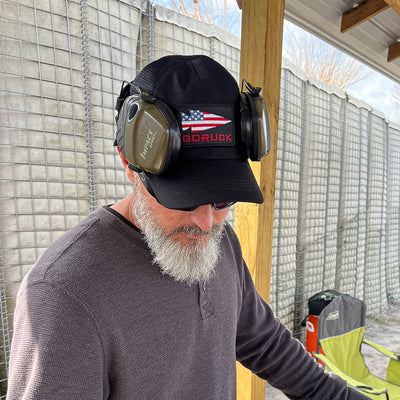 A man with a beard sporting a Cadre Hat - CORDURA® and earmuffs stands near a 500D CORDURA® wire mesh wall outdoors, channeling the rugged spirit of a Special Forces Cadre.