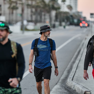 Sporting a blue shirt and black shorts, the individual confidently strolls down the city street. A military-style backpack strap is slung over his shoulder, and a Boonie Hat - Tactical - ToughDry tilts slightly over his eyes, giving him an air of adventure.