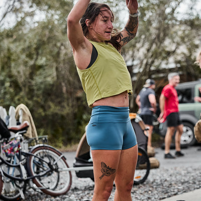 Exercising outdoors in a green crop top and blue Women’s Squat Shorts made with ToughFlex Fabric, this person is surrounded by bikes and people, shaded by trees.