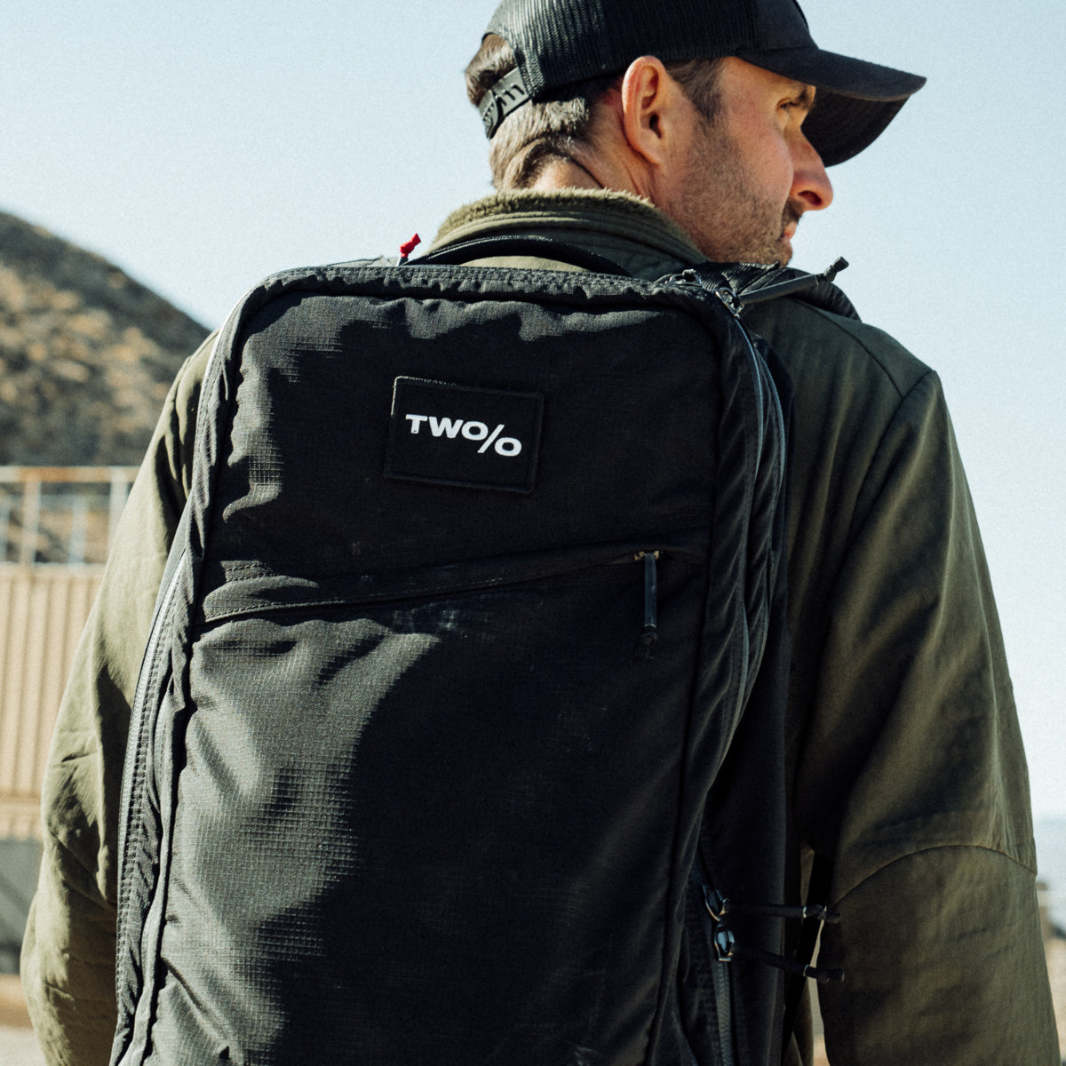 Under the clear sky, a man in a cap with a black backpack looks away. His GR2 - Ripstop ROBIC® - Michael Easter, made from durable 420D ROBIC Ripstop Nylon, hints at adventures supported by the Scars Lifetime Guarantee.