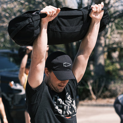 Donned in a Performance Running Hat - ToughMesh and a black T-shirt, the individual effortlessly hoists a large sandbag overhead outdoors.