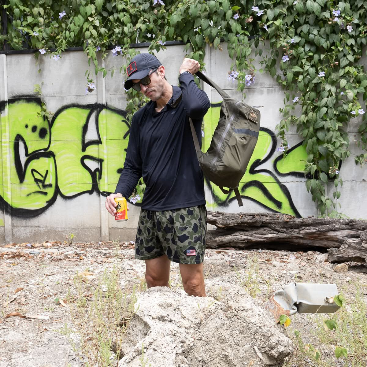 A person in a dark shirt and camo shorts stands by a graffiti-covered wall, holding a can. Their Packable Bullet Ruck - Ripstop ROBIC® backpack hints at adventure among the plants.