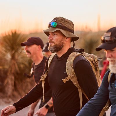 Three men hiking at sunset wear military-style gear with backpacks and Boonie Hats made of ToughDry® fabric, set against a blurred tree and sky background.