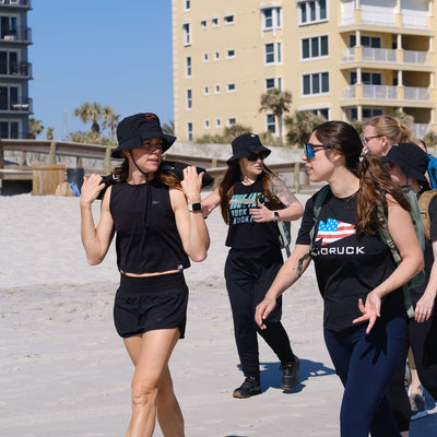 A group of women in sleek black exercise gear, with backpacks and Boonie Hat - Slick - ToughDry, stroll confidently along the sandy beach, ready for anything with protection from the elements thanks to their innovative apparel.