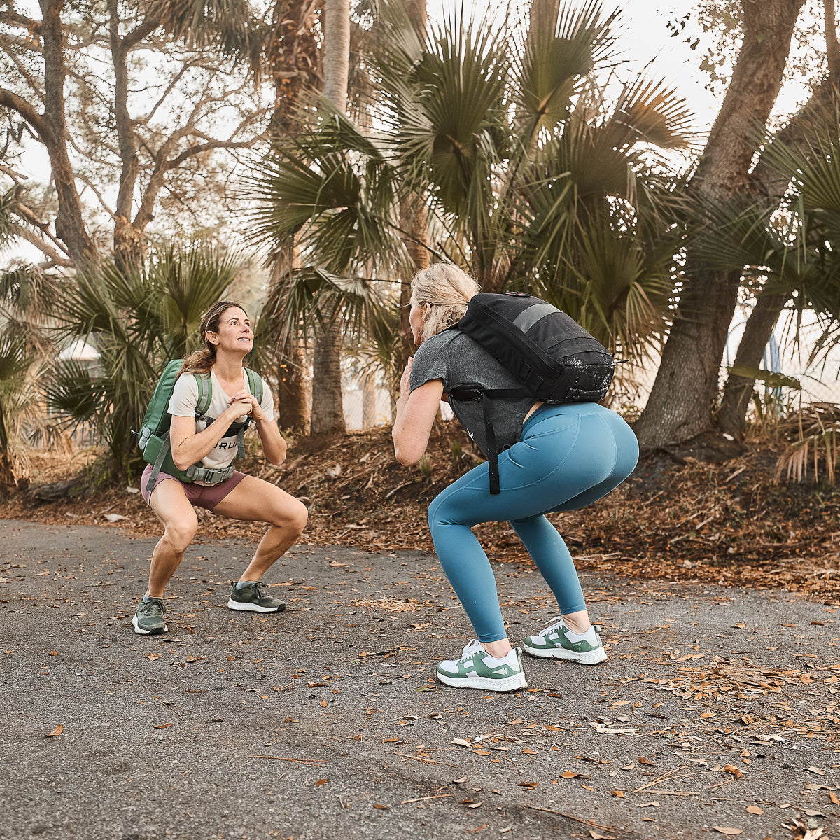 Two women are exercising outdoors, performing squats on a path with palm trees. They exemplify the durability and comfort of their Women’s Training Leggings Pocket - ToughFlex, which ensures resilience with every move.