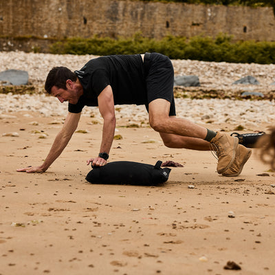 A man in a black T-shirt and shorts, wearing GORUCK's MACV-1 Hi-Speed - Mid Top rucking boots, exercises on a sandy beach. He is mid-movement with both hands and one knee touching the ground near a lightweight black sandbag. Rocky terrain and greenery are in the background.