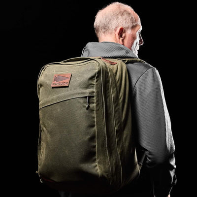 A man with short hair is wearing a dark jacket while carrying the GR2 - Waxed Canvas rucksack in olive green by GORUCK. This rucksack, made from waxed duck canvas, is adorned with a brown logo patch. The entirely black background accentuates the subject.