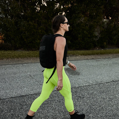 A person in neon green Women's Cropped Training Leggings Pocket - ToughFlex from GORUCK strolls along the road, sporting a black backpack and sunglasses.