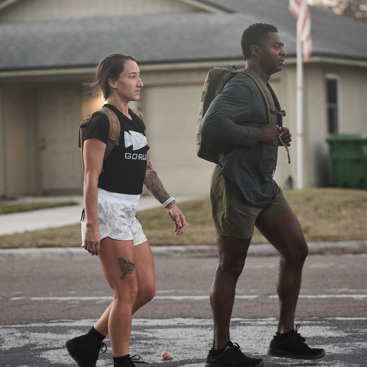 Two individuals clad in casual athletic attire, including GORUCK Women’s Training Shorts - ToughStretch, stroll past residential homes, both carrying backpacks as they walk outdoors.