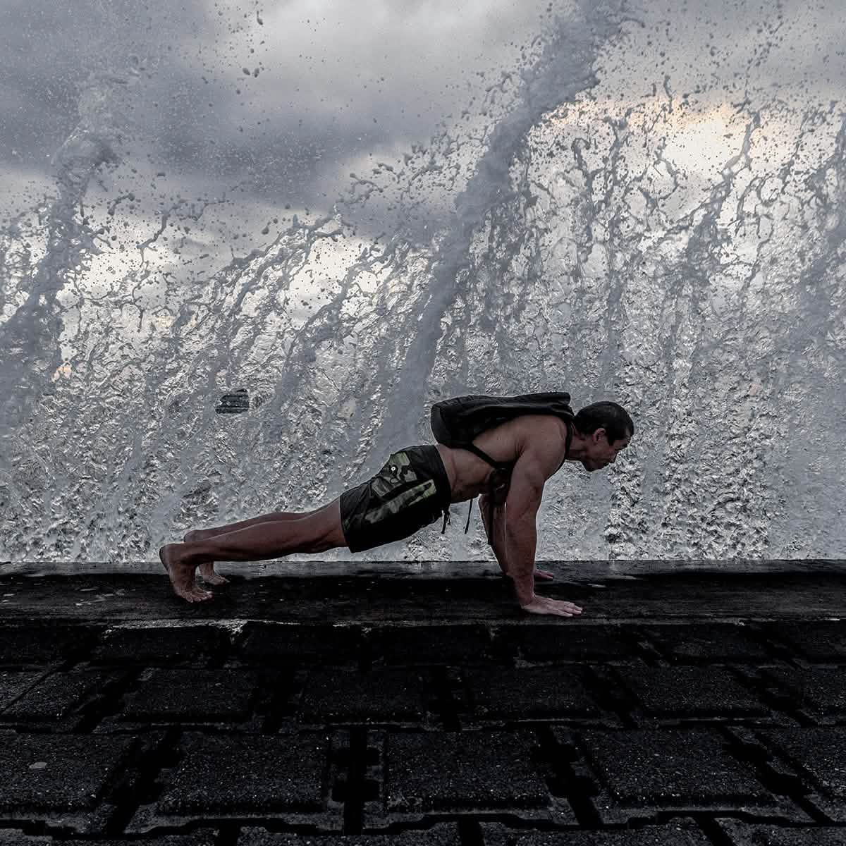 A person performs a push-up on a wet, textured ground with a large wave crashing in the background, creating a dramatic splash under a cloudy sky. They're sporting GORUCK's 300 Boardshorts and carrying a backpack, ready for adventure.