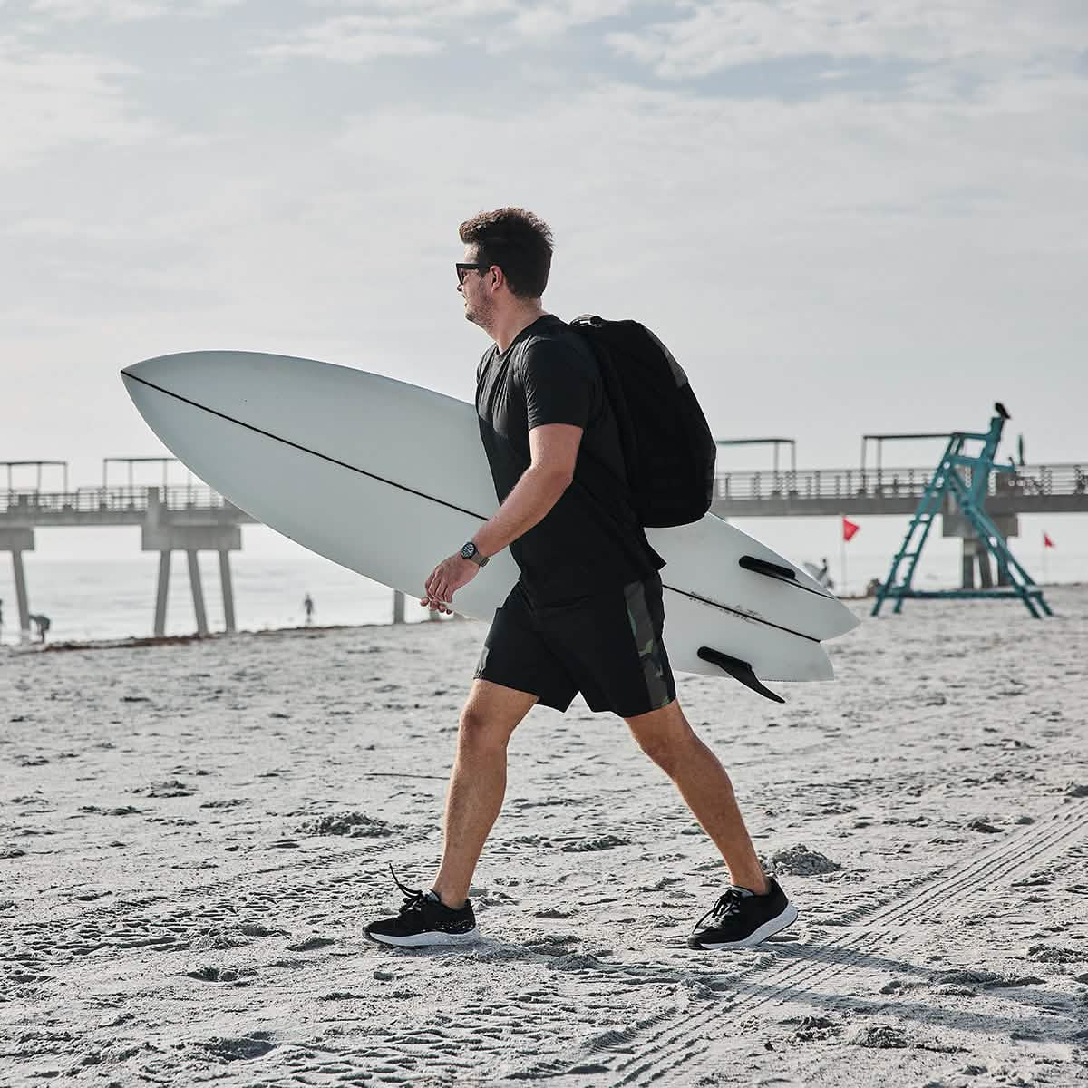 A man in a black t-shirt and GORUCK's 300 Boardshorts saunters along the sandy beach, carrying a white surfboard. With a backpack and sunglasses, he exudes coolness. In the background, people gather near a pier and lifeguard chair under the partly cloudy sky.