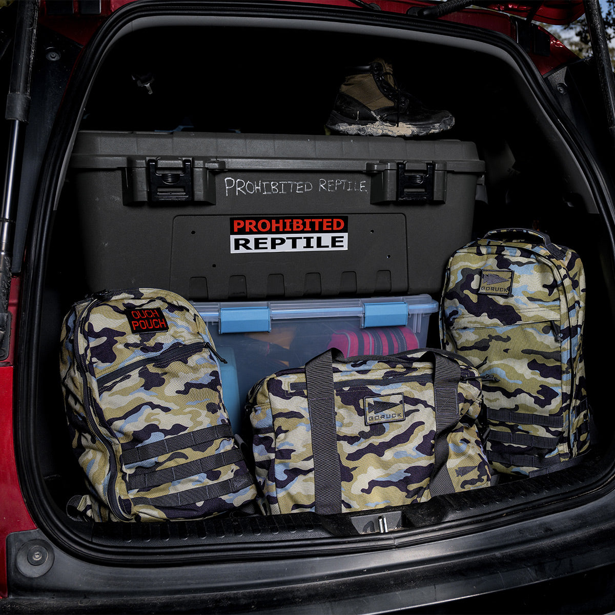 In the car trunk, camo bags were placed beside a container marked "Prohibited Reptile," and in the corner was a GORUCK Bullet Ruck Classic - Cordura, featuring a streamlined design and padded straps.