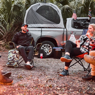 People sitting around a campfire, with the GORUCK GR1 USA - Cordura rucksack nearby and tents in the background, are surrounded by trees. A truck looms behind, reminiscent of Special Forces gatherings.