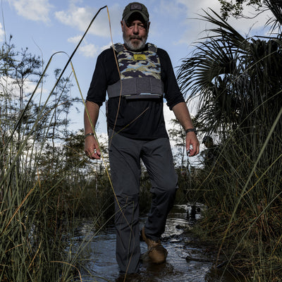 A man wearing outdoor gear and a camo vest strides through a marshy area with tall grass, his GORUCK Training Weight Vest 2.0 enhancing the workout. The sky is partly cloudy, and lush greenery envelops the scene.