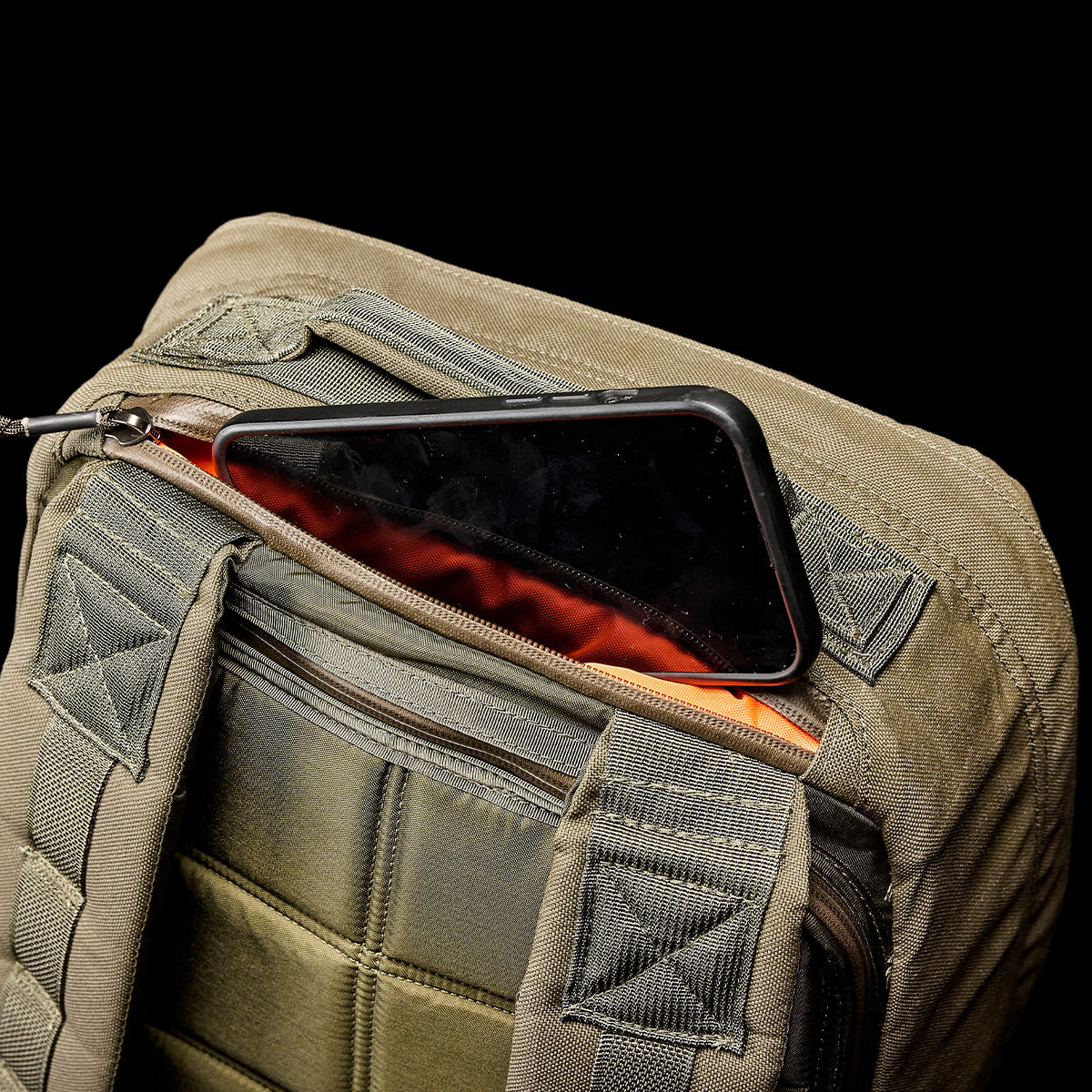 A smartphone peeks out from a GR1 USA - Cordura (The Original Ruck) backpack in khaki green, built with Special Forces-grade materials, against a black background.