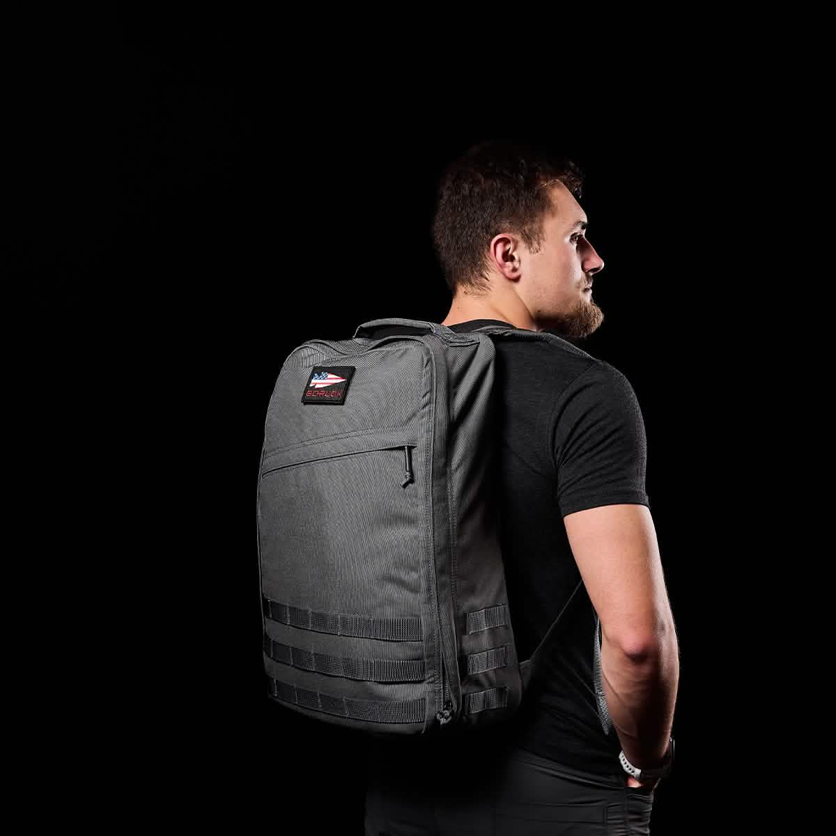 Man with a gray GR1 USA - Cordura (The Original Ruck) by GORUCK over one shoulder, featuring a bombproof laptop compartment, against a black background.