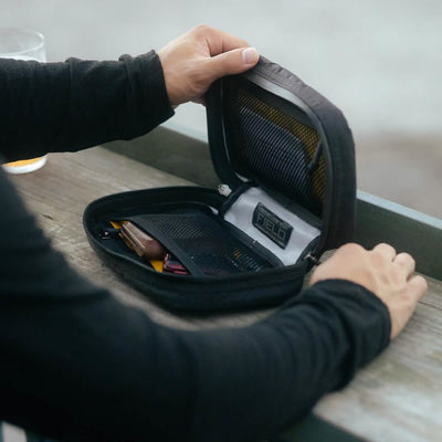 An individual in a long-sleeved black shirt opens a GR1 Field Pocket USA - X-PAC, produced by GORUCK, on a wooden bench. Inside the black zippered pouch, various items such as pens and small tools are neatly organized in mesh pockets.