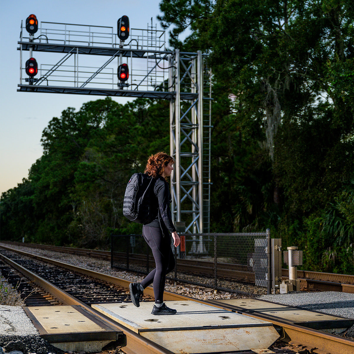 A person carrying the GORUCK GR2 - Dyneema backpack walks across a train track at dusk, with signal lights glowing softly above and casting shadows over the wooded surroundings.