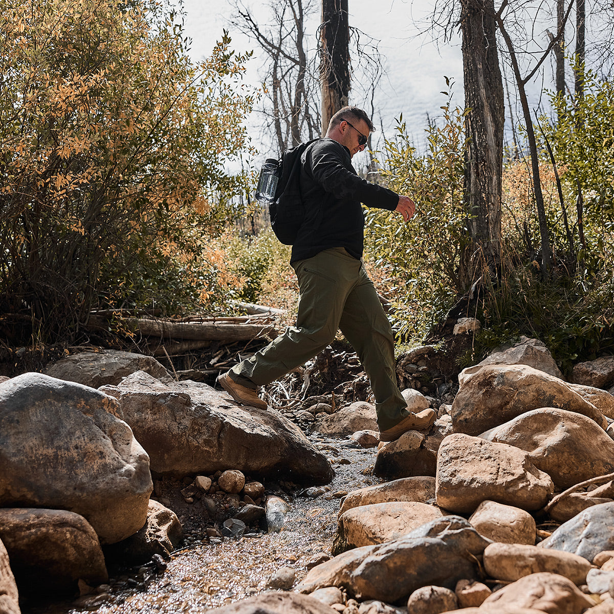 A man wearing a backpack steps confidently on rocks, crossing a stream in a wooded area, his Men’s Half Zip - Polartec Grid Fleece ensuring he stays warm and dry amidst the rugged terrain.