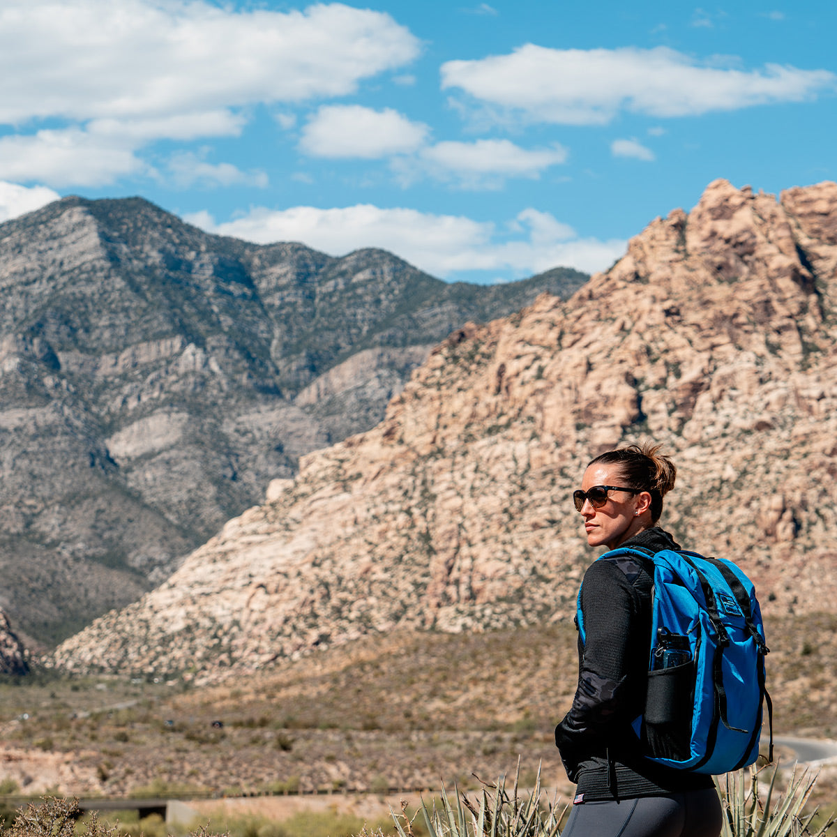 A woman wearing the Women’s Full Zip - PolarTec Grid Fleece from GORUCK hikes through a mountainous landscape under a partly cloudy sky, embracing the performance efficiency needed for such adventures.