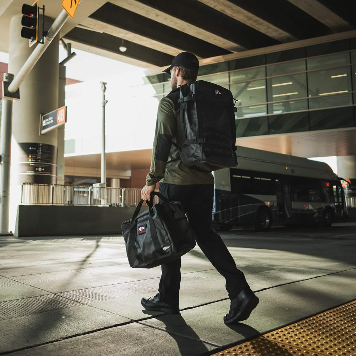 A person in dark clothing and a cap walks along a sidewalk near a bus station, carrying a GORUCK Kit Bag - Dyneema and an additional backpack. A bus is parked in the background under a multi-level structure with large glass windows.