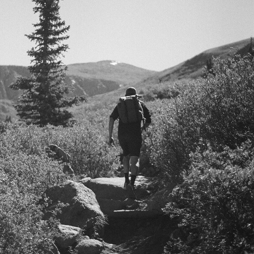 A hiker with a GORUCK M23 - Dyneema rucksack strides along a rocky trail, flanked by shrubs and mountains under a clear sky. A lone evergreen tree stands on the left. The image is in black and white, capturing the timeless spirit of adventure.