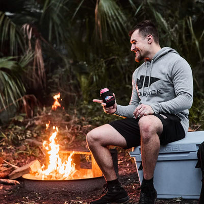 A man wearing a GORUCK Pullover - Poly-Blend and black shorts sits on a cooler, smiling and holding a drink. He is pointing towards a campfire surrounded by palm trees and greenery, capturing the adventurous spirit reminiscent of Special Forces roots.