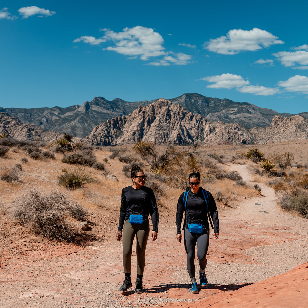 Two women with Bullet Rucks from GORUCK stroll along a dirt road, their gear packed efficiently for the journey.