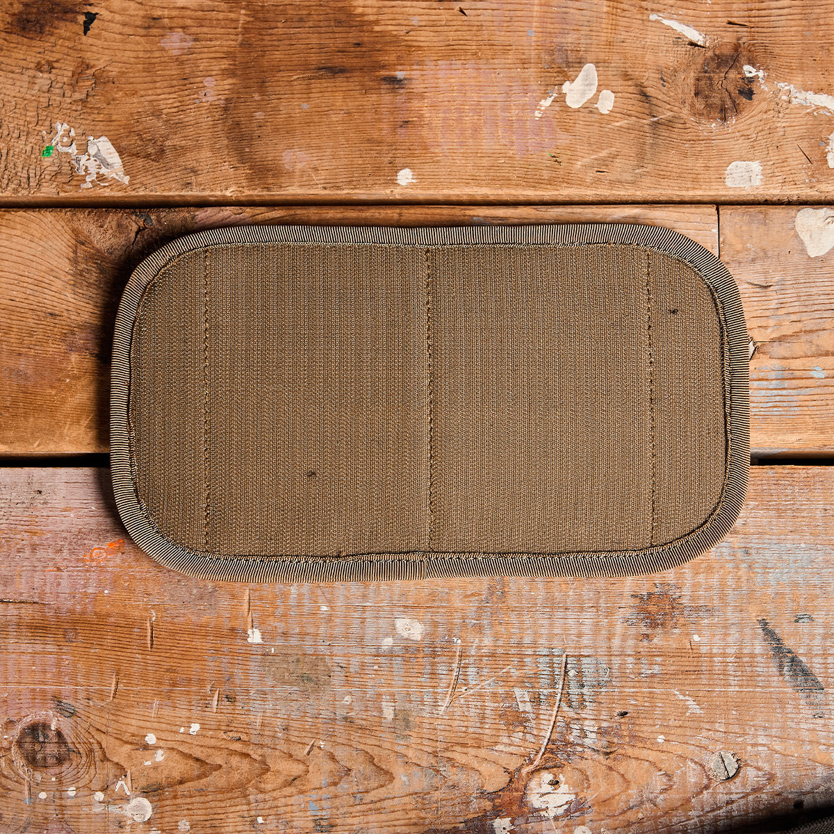 A rectangular tan woven fabric Admin Panel by GORUCK, featuring two vertical seams, rests on a rustic wooden surface speckled with faint paint splatters—an ideal companion for your EDC adventures.