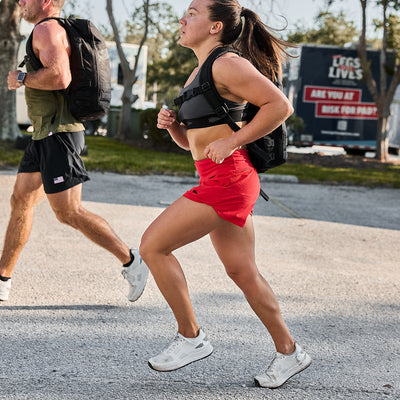 Two people are jogging outdoors on a sunny day. The woman in front is wearing a sports bra, red GORUCK Women’s Training Shorts made with ToughStretch fabric, and white sneakers. The man behind is in a tank top and black shorts. Both carry backpacks, with trees and a trailer in the background.