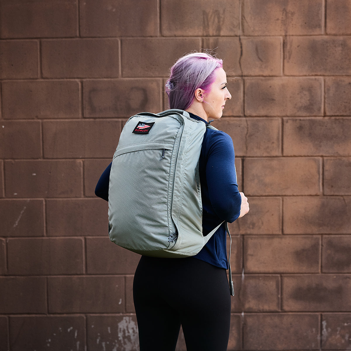 A person with purple hair, sporting an ultra-lightweight GR1 USA - Ripstop ROBIC® gray rucksack by GORUCK, stands against a brown brick wall.