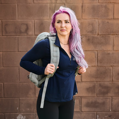 A person with long pink and purple hair, wearing a navy top and carrying the ultra lightweight, water-resistant GR1 USA Ripstop ROBIC® rucksack from GORUCK, stands in front of a brick wall.