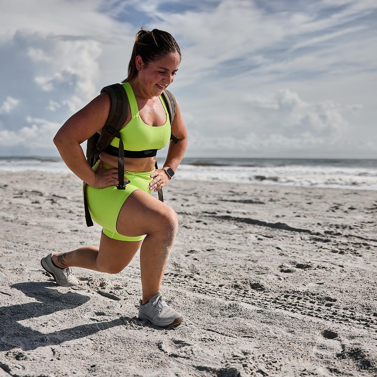 A person dressed in GORUCK Women's Biker Shorts made with ToughFlex fabric performs lunges on a sandy beach under a partly cloudy sky, sporting a backpack and smiling while waves crash in the background.