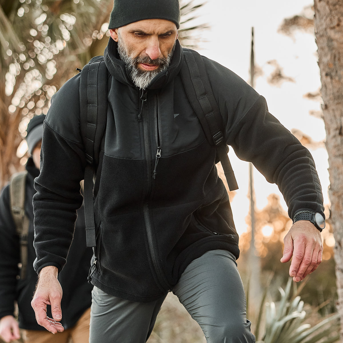 A man hikes uphill wearing a Men’s Task Force Dagger Jacket - Polartec Fleece and a beanie, reminiscent of Special Forces attire. He carries a backpack and wears a smartwatch, with trees in the background.