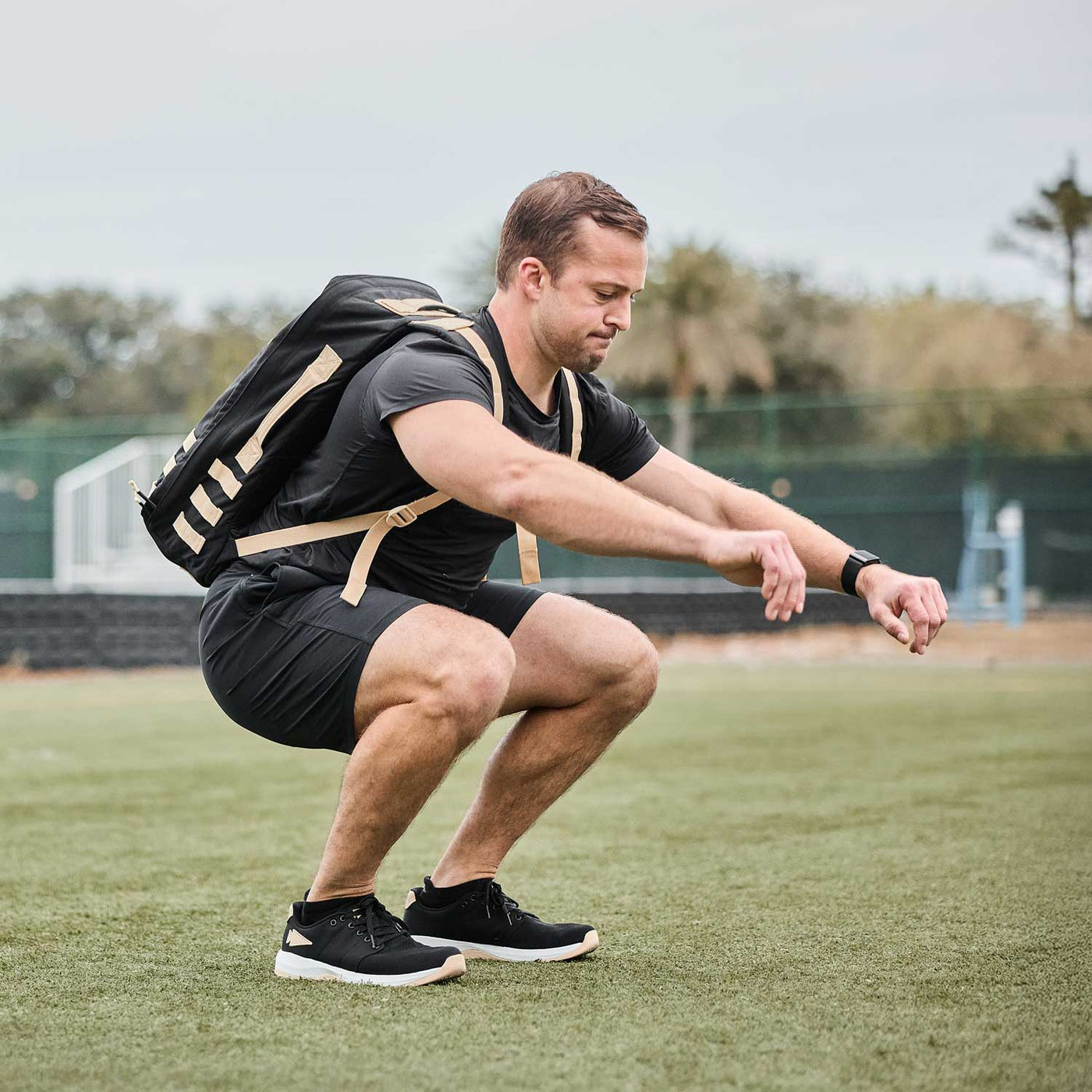 A man is doing squats outdoors on a field, wearing a backpack and fitness attire. His workout shoes are the Men’s Ballistic Trainers by GORUCK, designed for performance and featuring CORDURA® Ballistic Nylon for durability, in black, white, and gold with a gold reflective spearhead.