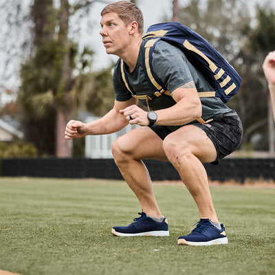 A man wearing a gray shirt and shorts performs squats on the grass, relying on his backpack crafted from durable CORDURA® Ballistic Nylon. For comfort and stability, he turns to GORUCK's Men’s Ballistic Trainers in Navy + White + Gold with a Gold Reflective Spearhead, ensuring 3X Support during his workout.