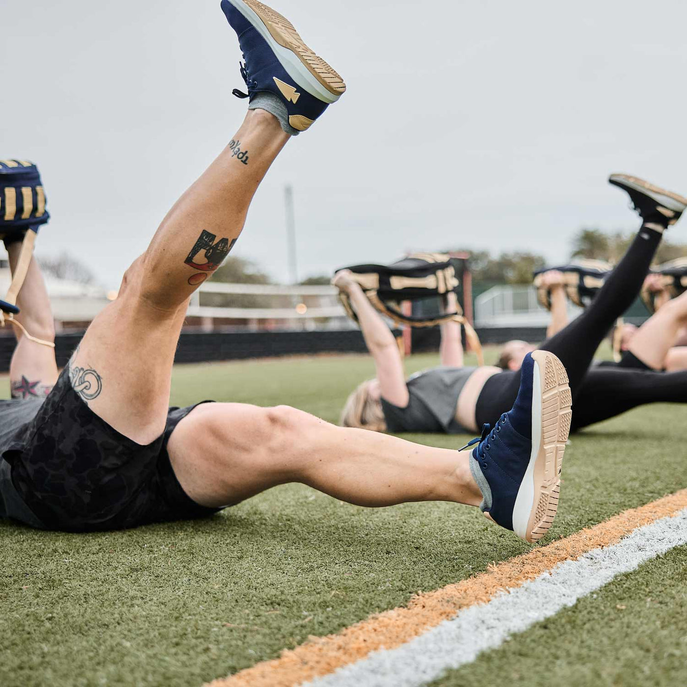 A group of people participating in functional fitness, lying on the grass and lifting weights with their legs raised, demonstrates versatility and strength-training benefits. Their GORUCK Men’s Ballistic Trainers in Navy + White + Gold with a Gold Reflective Spearhead provide stability and comfort as they power through their outdoor exercise routine.