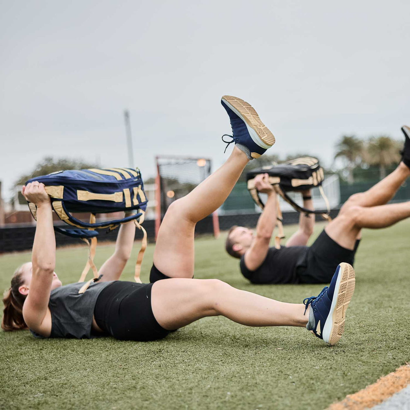 Two individuals exercising outdoors, lying on their backs and lifting weights with their legs elevated, demonstrate how crucial GORUCK's Women's Ballistic Trainers in Navy + White + Gold with a Gold Reflective Spearhead are for delivering the necessary support and comfort to achieve optimal performance.