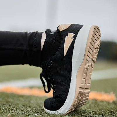 Close-up of a person wearing GORUCK Women's Ballistic Trainers in Black, White, and Gold with a Gold Reflective Spearhead on a grassy field, showcasing their durability as perfect functional fitness footwear.