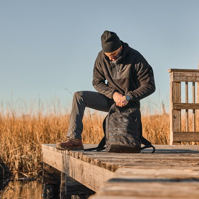 A man on a wooden dock searches his backpack, donned in the Men’s Task Force Dagger Jacket - Polartec Fleece. Tall grass sways behind him, evoking rugged adventures.