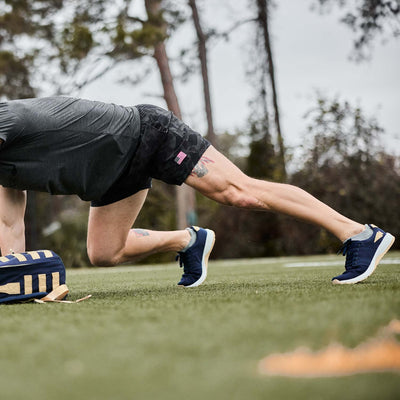 A person in navy and white trainers with gold reflective details from GORUCK performs a push-up on the grass, with their backpack made of durable CORDURA® Ballistic Nylon resting nearby.