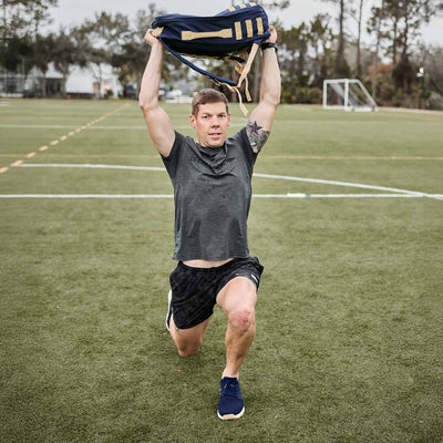 A man performs lunges on a sports field, lifting a backpack overhead with both hands, showcasing the durability of GORUCK’s Men’s Ballistic Trainers in Navy, White, and Gold with a Gold Reflective Spearhead.