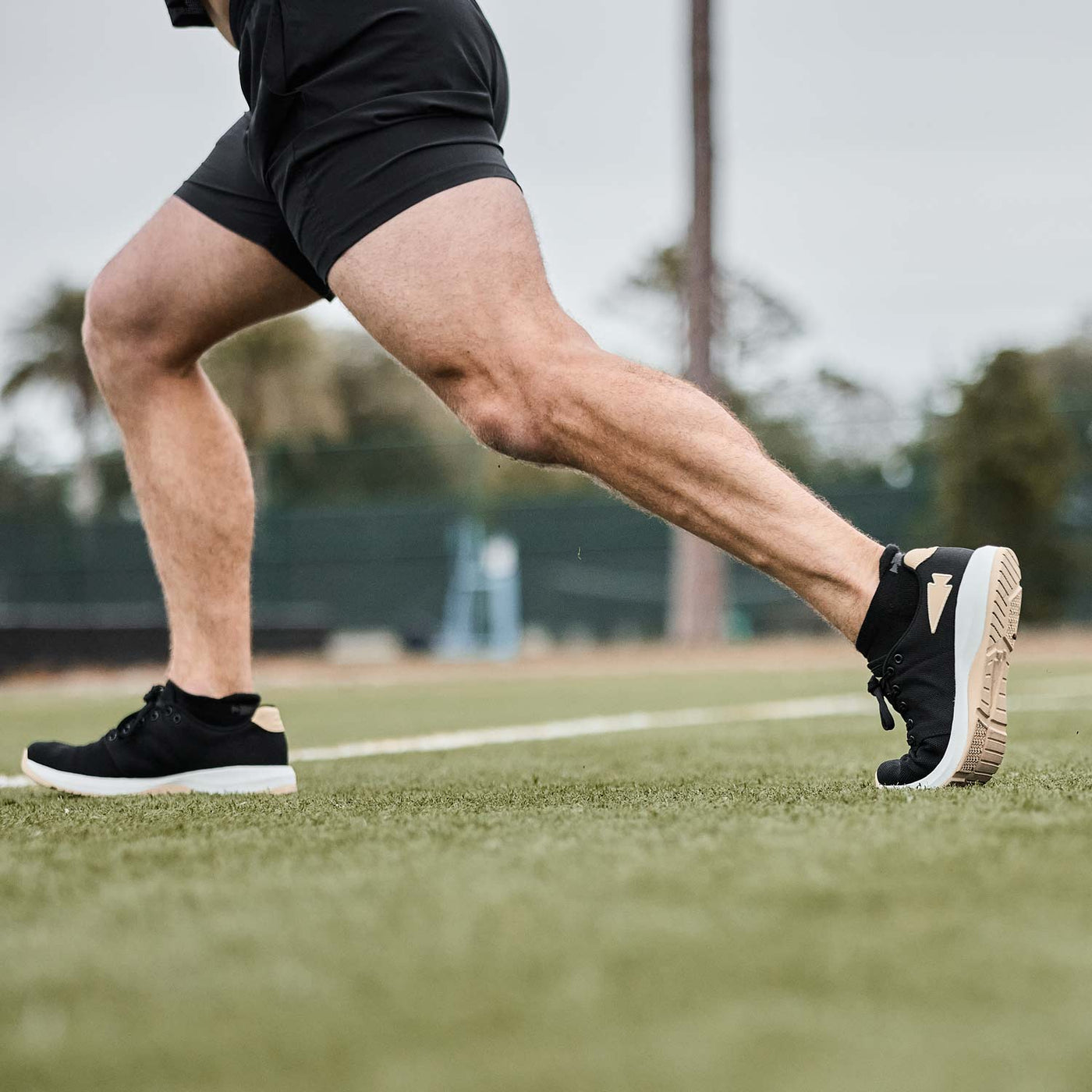 A person is running on grass, showcasing the durability of black athletic workout shoes and shorts. The GORUCK Men's Ballistic Trainers in Black, White, and Gold with a Gold Reflective Spearhead feature a sleek design crafted from CORDURA® Ballistic Nylon for ultimate endurance.