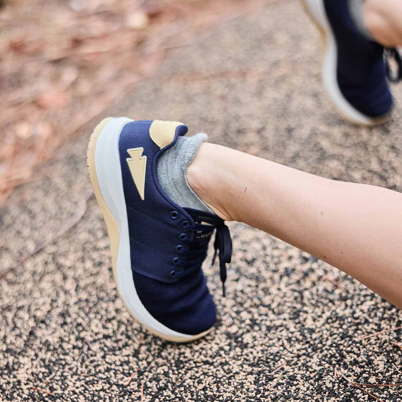 A person is walking on a textured pathway wearing GORUCK's Women's Ballistic Trainers in navy, white, and gold with a gold reflective spearhead, crafted from CORDURA® Ballistic Nylon.