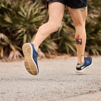 A person running on a path in GORUCK Men’s Ballistic Trainers, with navy and white shoes featuring gold accents, showcasing a visible calf tattoo. The trainers incorporate 3X Support construction and durable CORDURA® Ballistic Nylon, all against a backdrop of blurred greenery.