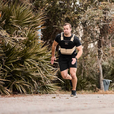 As a man jogs outdoors, sporting the GORUCK Men’s Ballistic Trainers in Black, White, and Gold with Gold Reflective Spearhead and a weighted vest, trees provide a scenic backdrop.