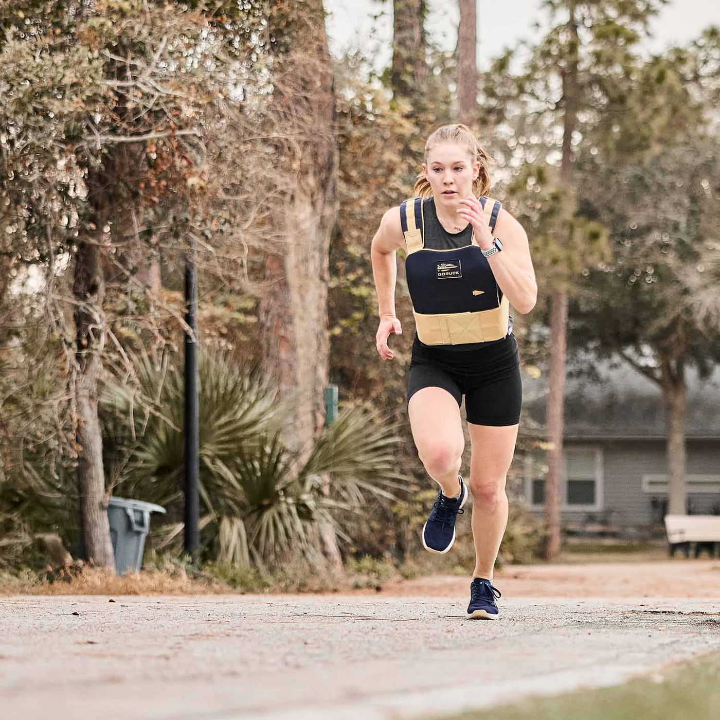 A woman wearing a weighted vest and GORUCK's Women's Ballistic Trainers in Navy, White, and Gold with a gold reflective spearhead sprints outdoors on a paved path surrounded by trees.