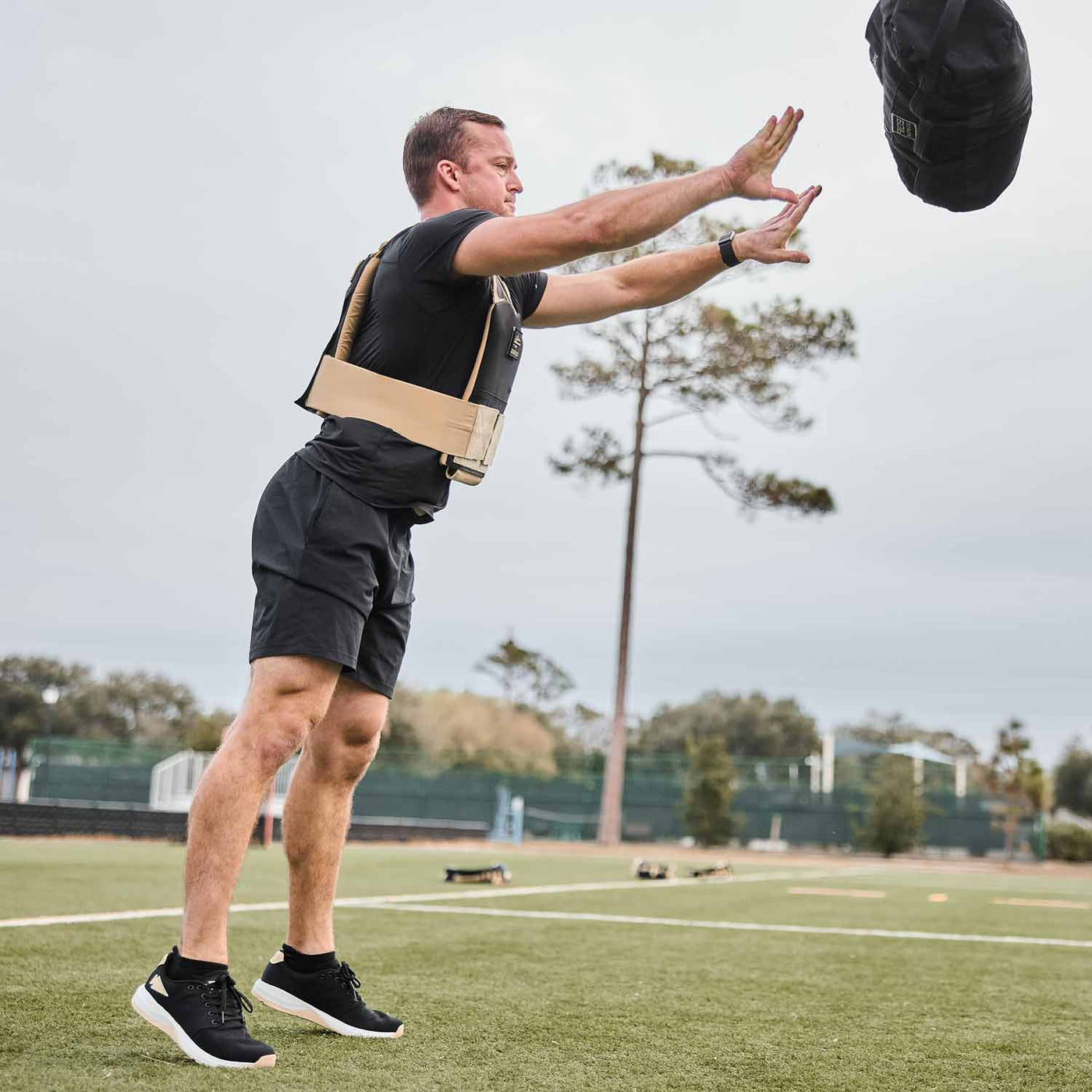 A man, donned in GORUCK's Men’s Ballistic Trainers in Black, White, and Gold with a Gold Reflective Spearhead, is hurling a sandbag across a grassy field.